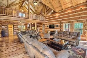 A spacious log cabin interior with a high ceiling, multiple seating areas, and a balcony overlooking the forest.