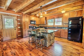 Une cuisine rustique avec des poutres en bois, un îlot central en pierre avec des tabourets de bar, un réfrigérateur noir et une fenêtre donnant sur les arbres.