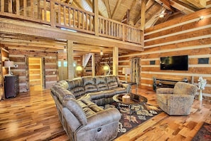 Ein Interieur einer Blockhütte mit einem großen Ledersofa, einem gemusterten Sessel, einem Flachbildfernseher und einer Holztreppe.