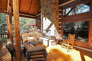 Une terrasse en bois avec des sièges d’extérieur, une cheminée en pierre et de grandes fenêtres donnant sur une zone boisée.