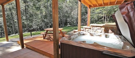A hot tub under a wooden pergola with a picnic table nearby, surrounded by a forested area.