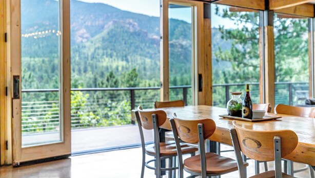 A dining area with a large table and chairs, a view of mountains and greenery through large windows, and a wooden ceiling with hanging lights. - Spectacular Views - Stunning Modern Broadmoor Home (Colorado Springs)