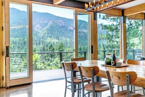 Een eetgedeelte met een grote tafel en stoelen, uitzicht op bergen en groen door grote ramen, en een houten plafond met hangende lampen.