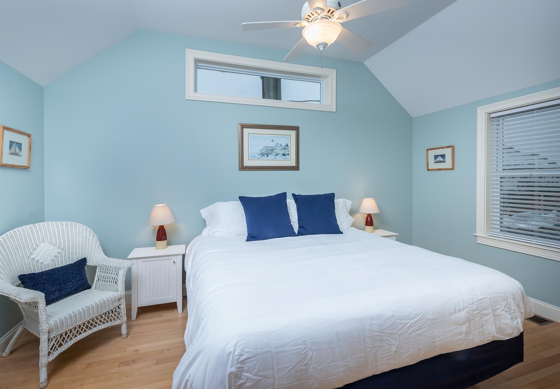 A bedroom with a bed, a white wicker chair, two bedside tables with lamps, a ceiling fan, and a window with blinds.