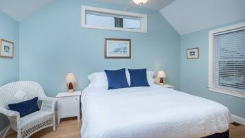 A bedroom with a bed, a white wicker chair, two bedside tables with lamps, a ceiling fan, and a window with blinds.