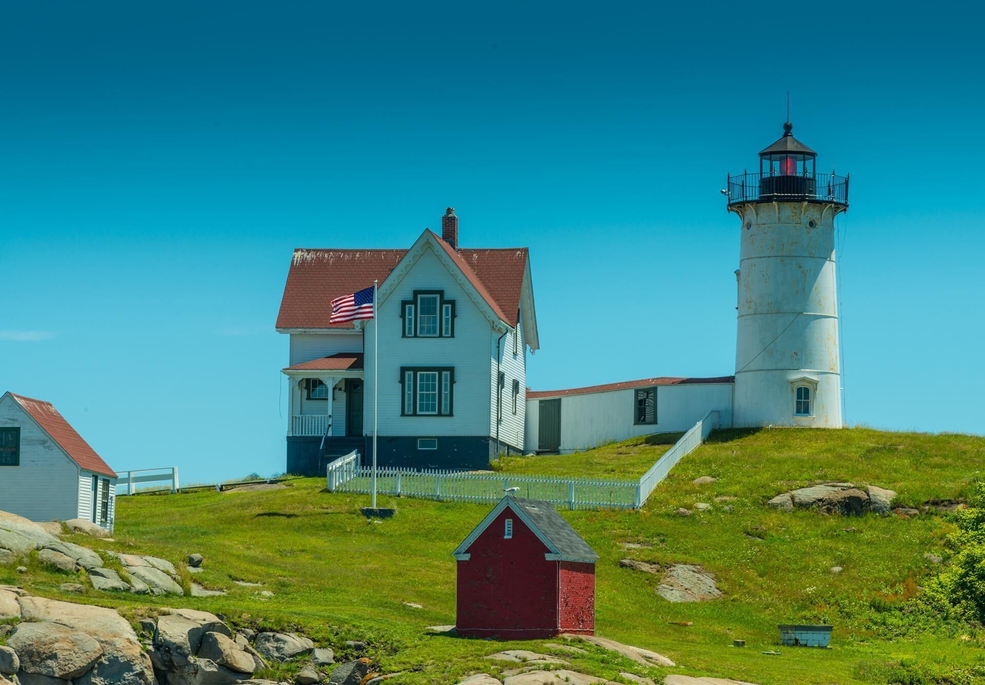 A lighthouse with a white exterior and a red roof, a white house with a red roof, and a small red shed.