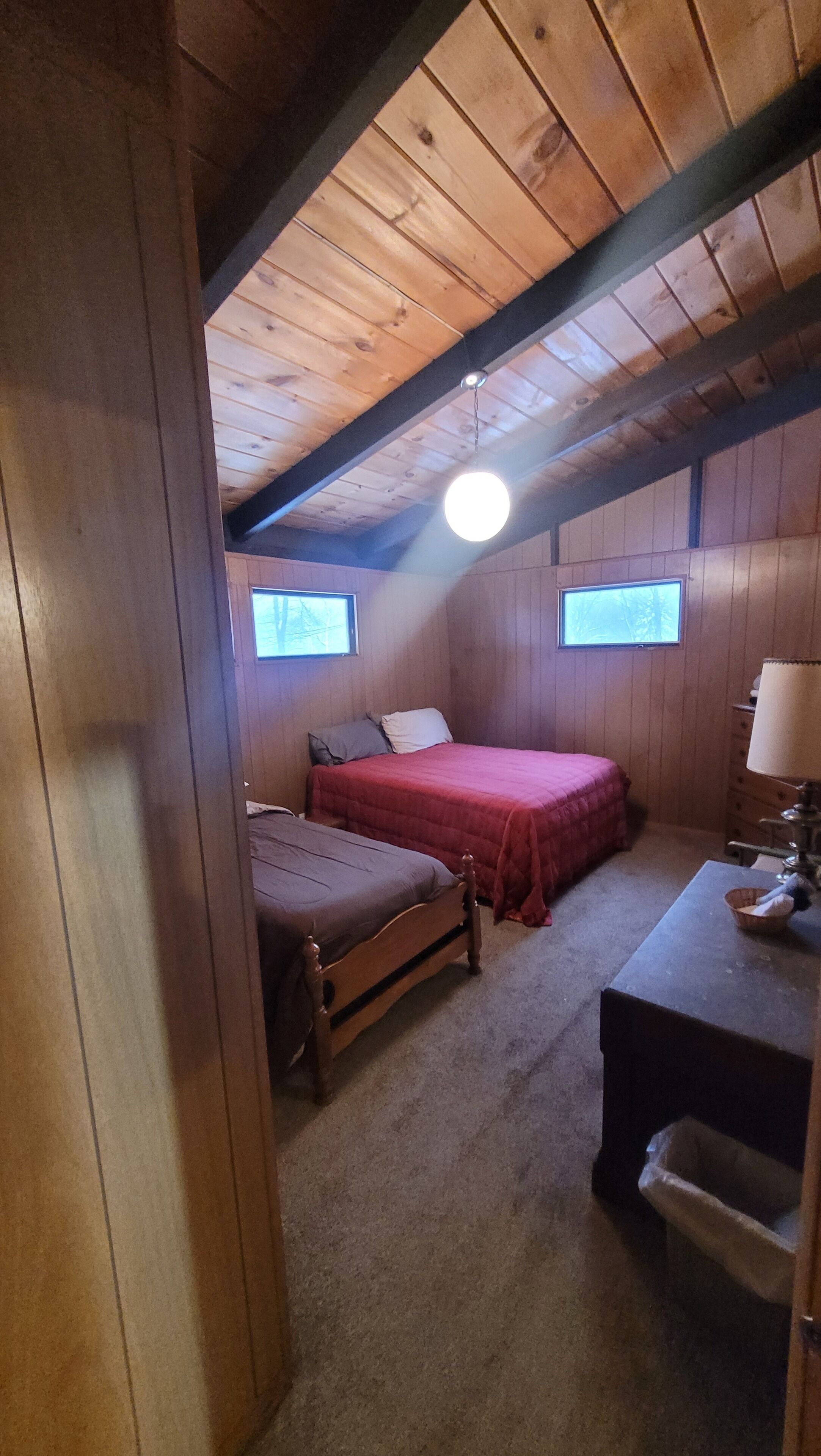 A small, cozy bedroom with wooden walls and a sloped ceiling. There are two beds, a desk, and a lamp.