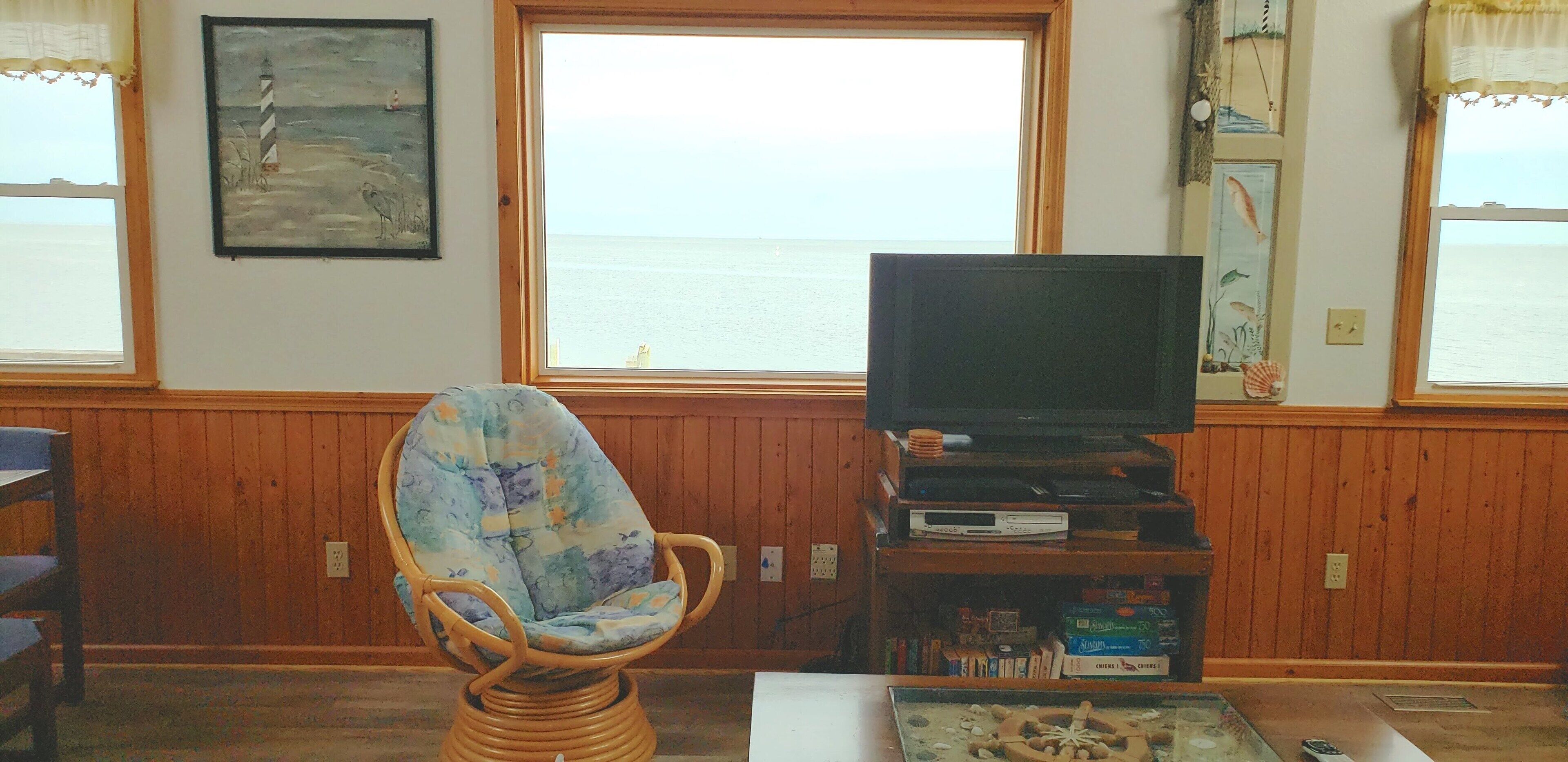 Een woonkamer met een rieten stoel, een flatscreen televisie op een houten standaard, en uitzicht op de zee door grote ramen.