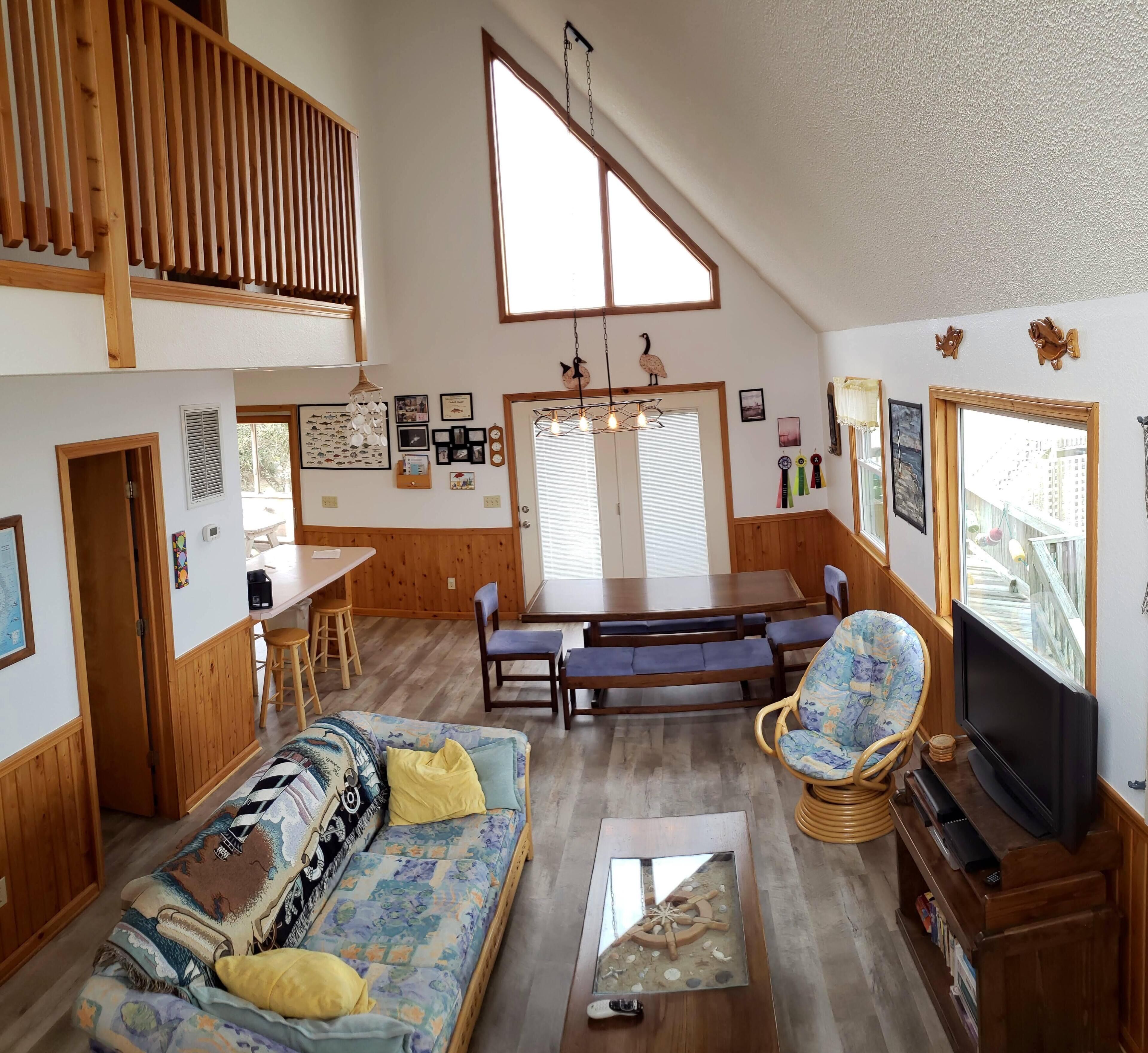 A spacious living area with a high ceiling, wooden beams, a dining table with chairs, a sofa with cushions, a television, and a bookshelf.