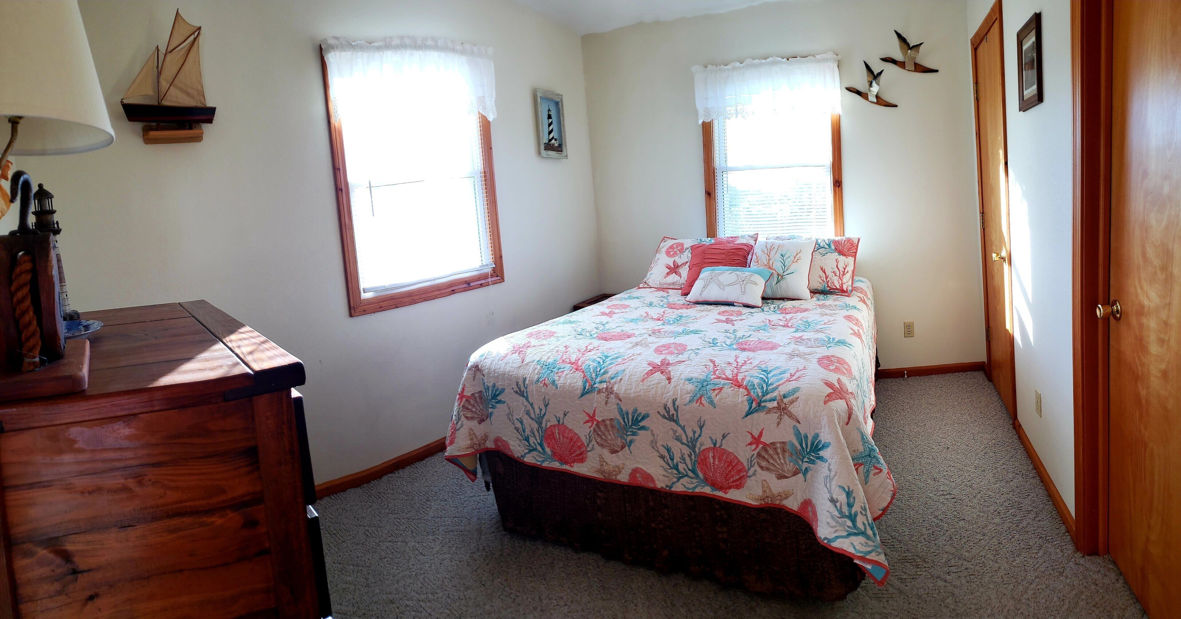 A bedroom with a bed, a dresser, two windows with curtains, a wall-mounted shelf with a ship model, and a door with a brass handle.