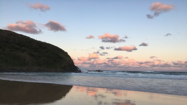 Ein Strand mit ruhigem Wasser, das den Himmel spiegelt, einem sandigen Ufer und einem grünen Hügel im Hintergrund.