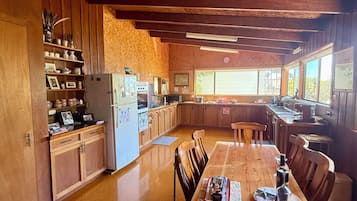 Cuina àmplia amb armaris de fusta, taula de dinar amb caires i un gran finestral que deixa entrar llum natural.