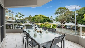 A balcony with a glass table and chairs, overlooking a street with parked cars and greenery.