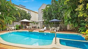 A swimming pool with lounge chairs and umbrellas, surrounded by lush greenery and a building with a white facade.