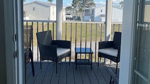 A covered porch with two wicker chairs and a small table, overlooking a residential area with houses and a tree.