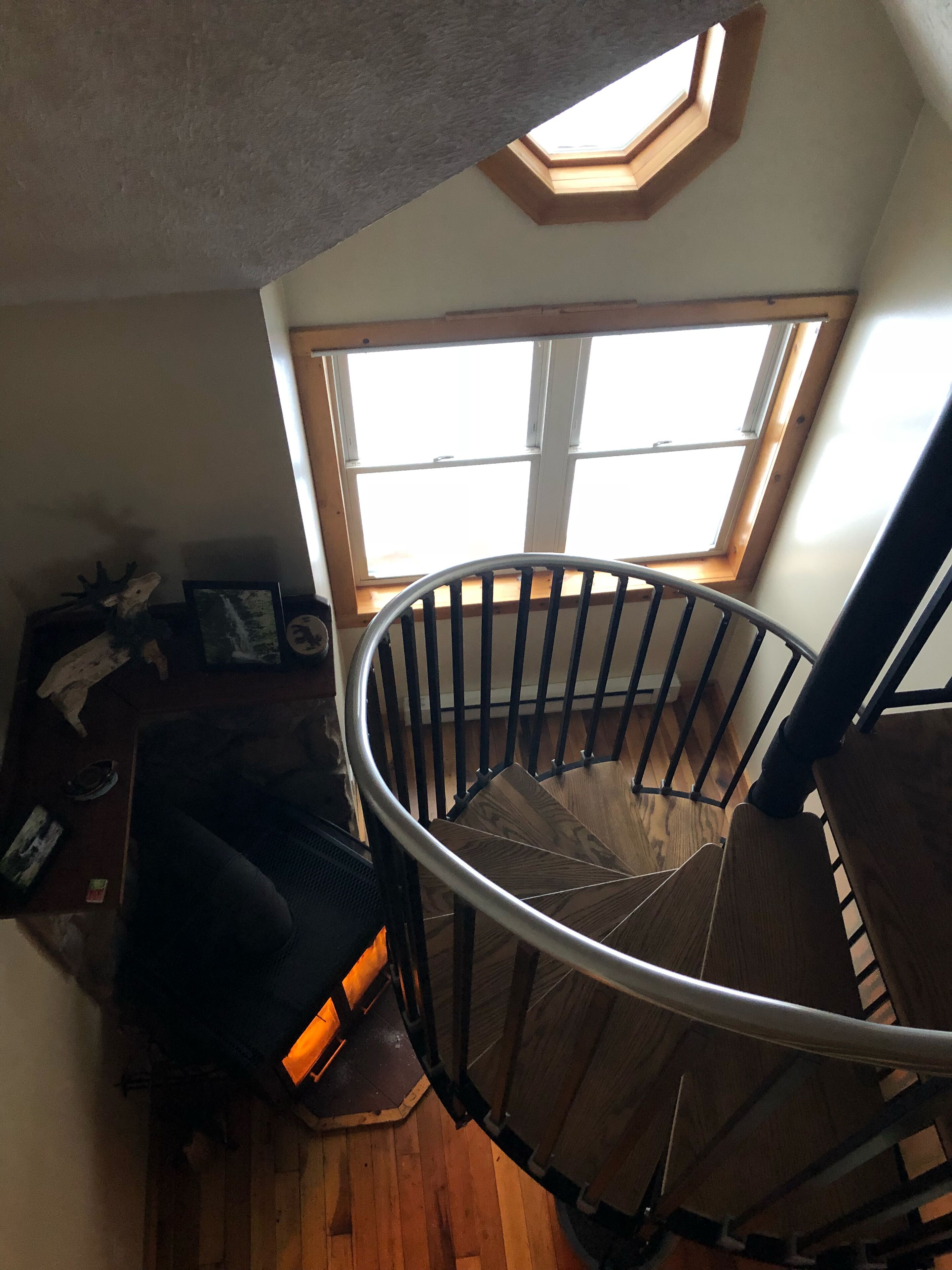 Una escalera de caracol con acabado de madera y barandillas metálicas, que conduce a una habitación con tragaluz y un ventanal grande.