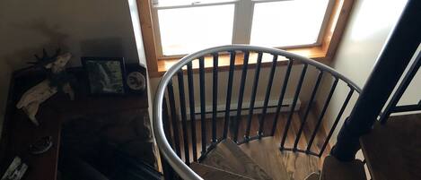 Una escalera de caracol con acabado de madera y barandillas metálicas, que conduce a una habitación con tragaluz y un ventanal grande.