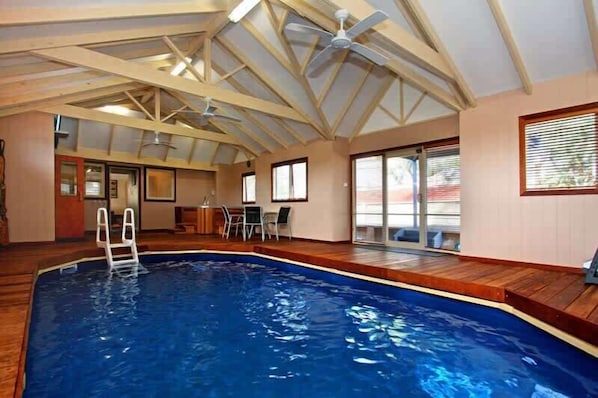 An indoor swimming pool with a wooden deck, a ladder, and a ceiling fan.