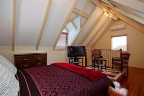 A bedroom with a sloped ceiling, a bed with a patterned bedspread, a television, and a seating area with chairs.