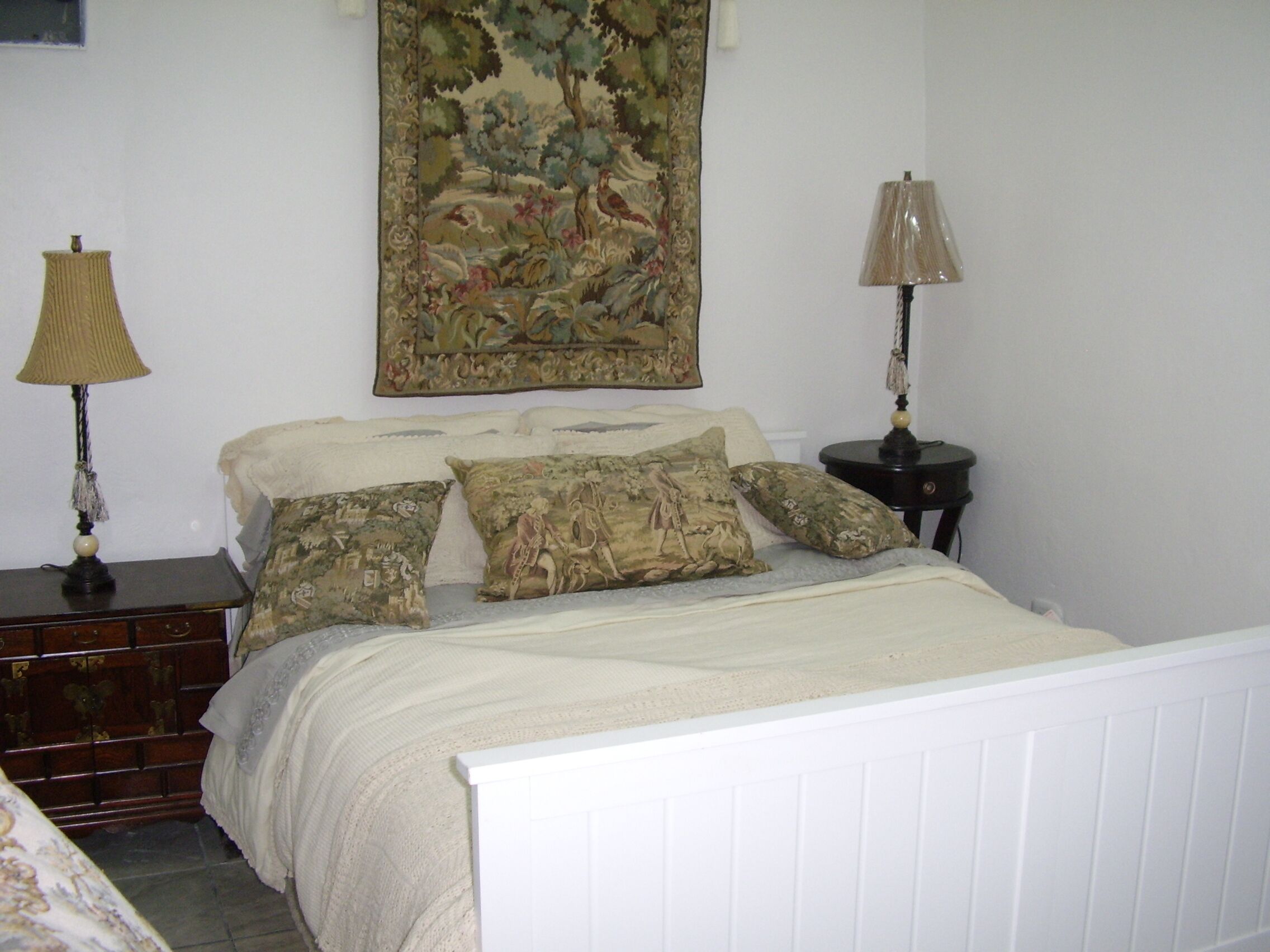 Un dormitorio con una cama, dos mesitas de noche, una lámpara, un aparador y un tapiz en la pared.