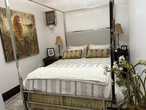 A four-poster bed with a white and striped bedspread, a nightstand with a lamp, a framed picture on the wall, and a painting of a parrot.