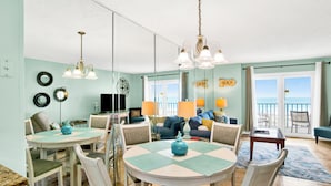 A dining area with a round table, chairs, and a blue vase centerpiece. There is a sofa and a television in the background. - Surfside Shores II 2204 Directly On The Gulf Of America (Gulf Shores)