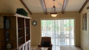 Un salón espacioso con techo alto, una estantería de madera, un sillón estampado y una zona de comedor con mesa y sillas.