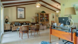 Un salón amplio con techo alto, vigas de madera, chimenea, estantería y escritorio con ordenador.