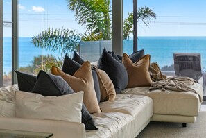 Un salón moderno con un amplio ventanal con vistas al océano, un sofá color beige con varios cojines y una mesa de centro de vidrio.
