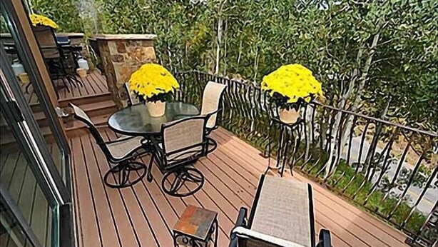 A wooden deck with a glass-top table and four chairs, surrounded by a metal railing and greenery. - Large 6 Bedroom Home- Ski-in / Ski-out Access (202983-2164) (Snowmass Village)