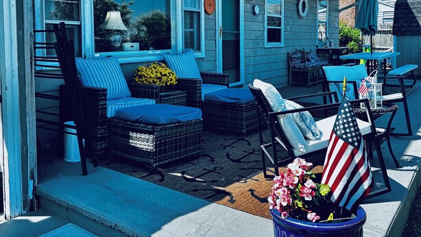 A covered porch with outdoor furniture, including wicker chairs and a sofa, and a potted plant with flowers. -  Beautiful Waterfront Home (Misquamicut)