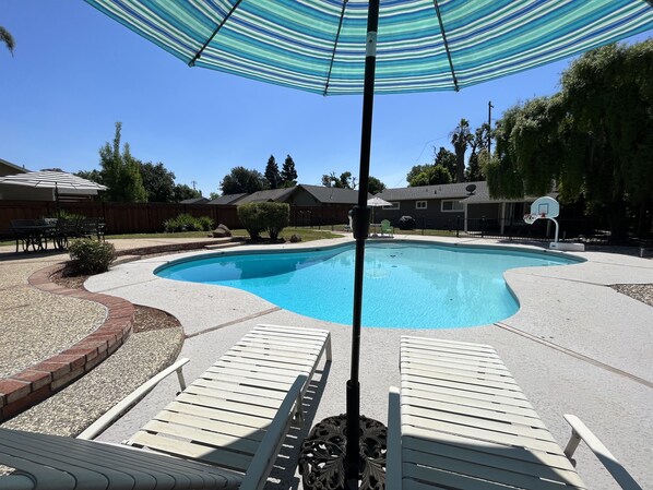 Un jardin avec une piscine, deux chaises longues blanches, un panier de basketball et un parasol à rayures.