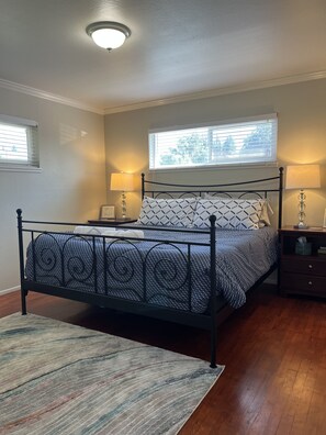 Une chambre à coucher avec un lit en métal, deux tables de chevet avec des lampes, une fenêtre avec des stores et un tapis à motifs.