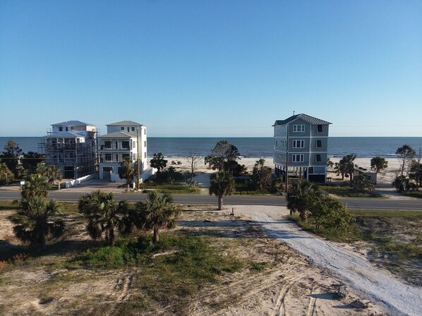 Two-story buildings with modern architecture stand amidst palm trees and a dirt road leading to the ocean.