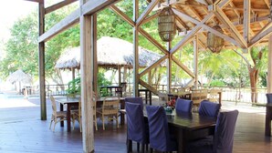 An outdoor wooden deck with tables and chairs, surrounded by thatched-roof structures and lush greenery. - Beachfront | Beachside Private Pool | Immaculately Kept | Private Dive Resort (Roatan)