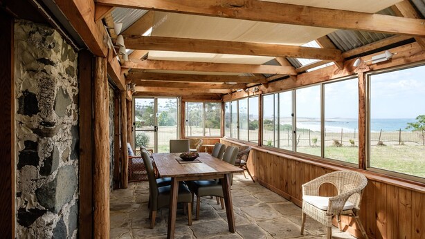 Ein überdachter Essbereich mit Steinwand, Holzbalken und großen Fenstern mit Blick auf einen Strand.
