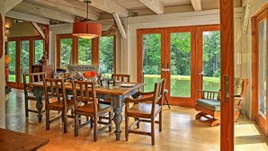 A spacious dining area with a wooden table and chairs, large windows offering a view of the outdoors, and a cozy seating area with a rocking chair. - House Belleayre peaceful spring water swimming pond (Fleischmanns)