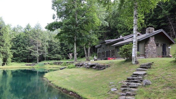 A lakeside lodge with a stone chimney, a wooden deck, and a stone pathway leading to the building. - House Belleayre peaceful spring water swimming pond (Fleischmanns)