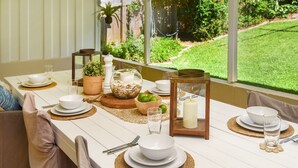 A dining table set with white plates, bowls, and glasses, surrounded by natural light and greenery. - Jetty Beach House (Coffs Harbour)