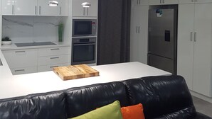A modern kitchen with white cabinets, a built-in oven, and a refrigerator. There is a black leather couch with colorful cushions in the foreground.