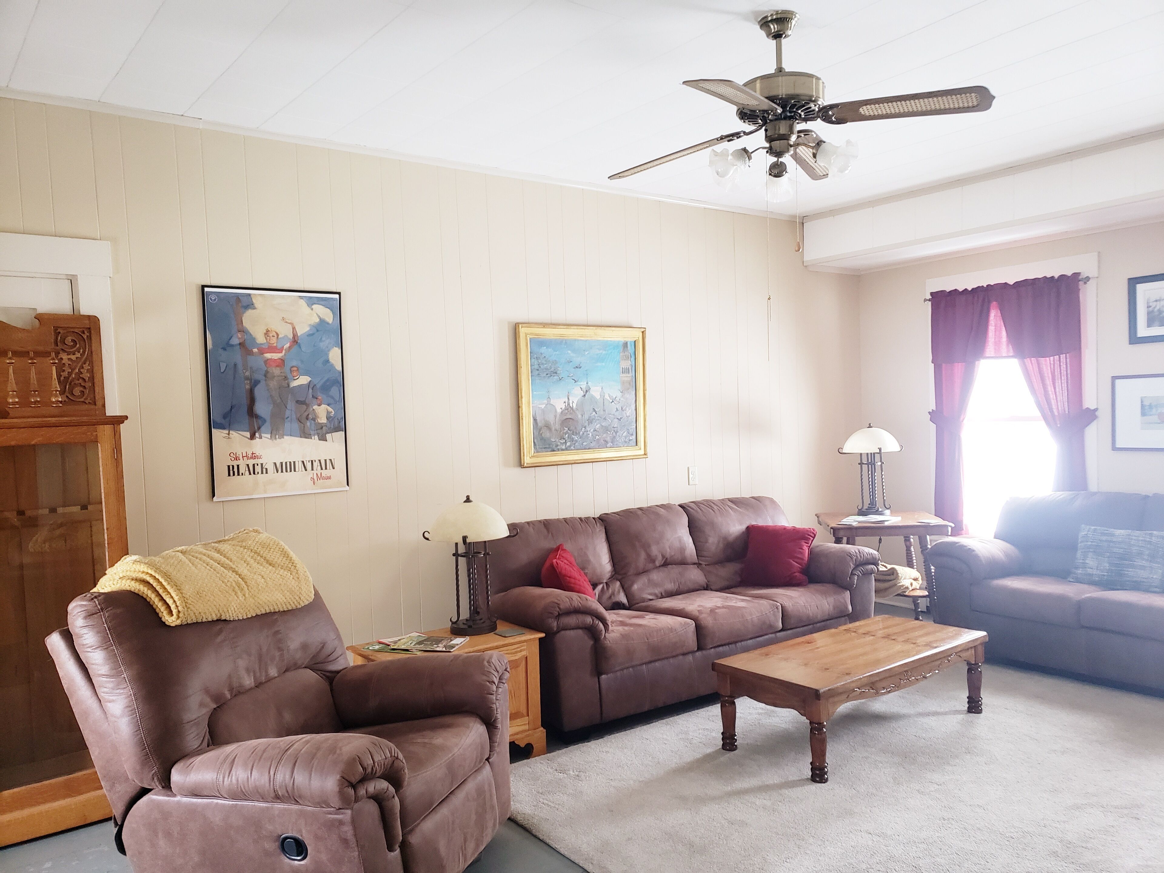 A living room with a brown leather sofa, a wooden coffee table, and a ceiling fan. There are framed pictures on the walls and a lamp on a side table.