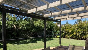 Un patio cubierto con una mesa y sillas, una pérgola con paneles solares y vistas a un césped y árboles bien cuidados.