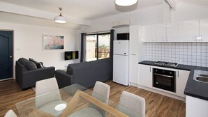 Una cocina moderna con armarios blancos, encimera negra y horno empotrado. Hay un comedor con mesa de vidrio y sillas.