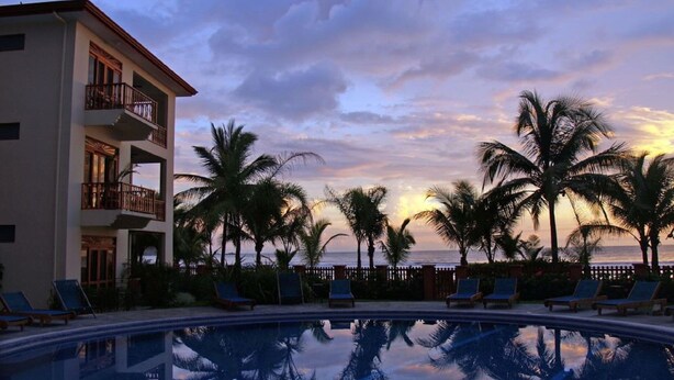 Une piscine d’un complexe hôtelier, bordée de palmiers et offrant une vue sur l’océan au coucher du soleil.