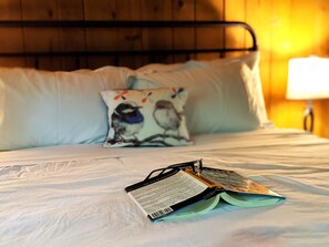 A bed with a pillow featuring a bird design, a book, and a lamp in a room with wooden paneling. - Amenities & Resort Access Included, Cabin 3 (Phelps)