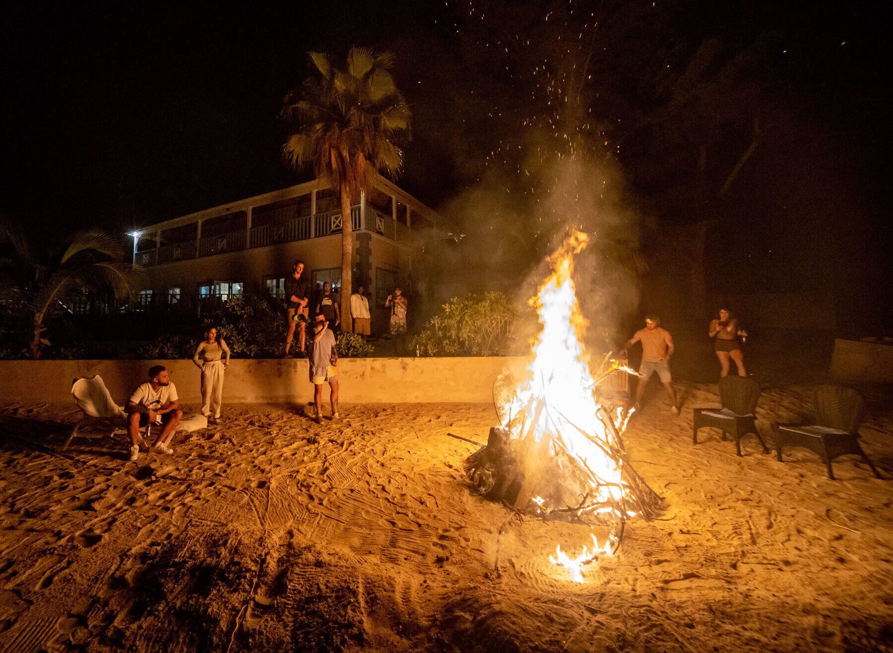 A beach scene with a campfire, people gathered around, and a building in the background.