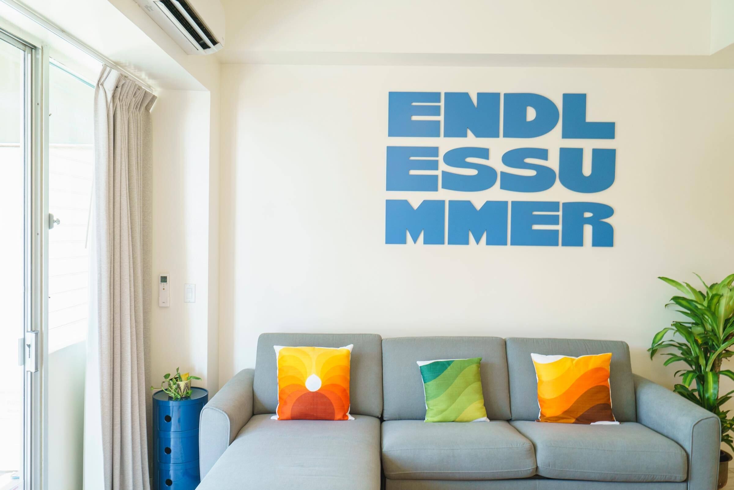 Un salon moderne avec un canapé gris, des coussins colorés et un mur orné de l’inscription « SUMMER ENDLESS ».