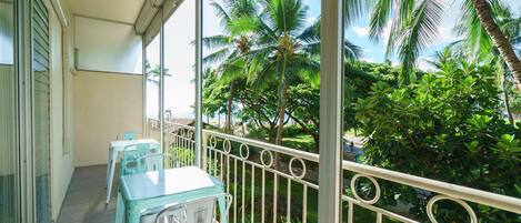 A balcony with a table and chairs, surrounded by a metal railing and lush greenery including palm trees.