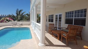 Piscina com deck revestido em azulejo, colunas brancas e área de varanda coberta com móveis de madeira e grandes janelas.