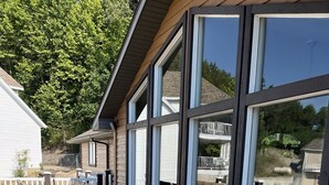Eine Holzterrasse mit Liegestuhl und Tisch, großen Fenstern und Blick auf ein Gebäude und Bäume.